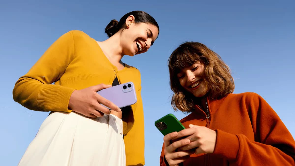 Dos mujeres sonríen con sus teléfonos en la mano contra un cielo azul